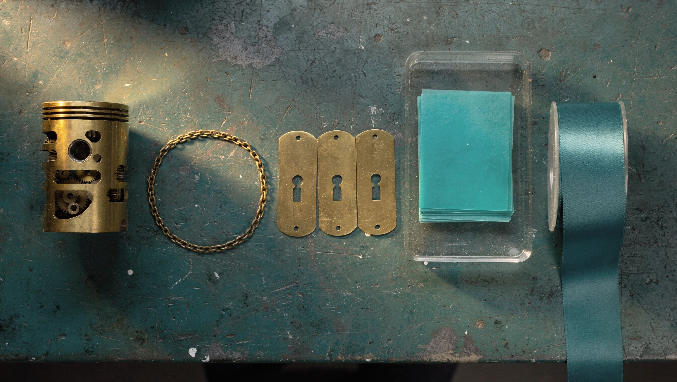 Five objects laid out in a row on a dark teal bench under one warm raking light — a small brass engine, a closed loop of fine chain, three brass plates with keyholes, a glass tray of stacked teal sheets, and a spool trailing a ribbon of tape off the edge of the frame.